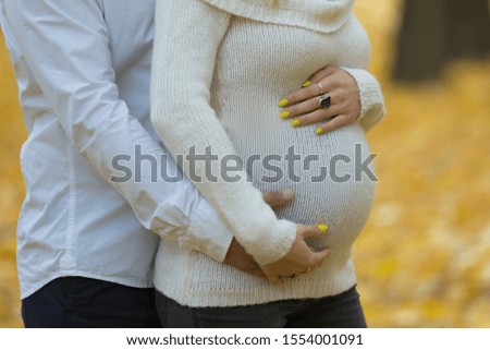 a man's arms hug the belly of a pregnant woman in a white knitted sweater in a Park in autumn against a background of yellow leaves