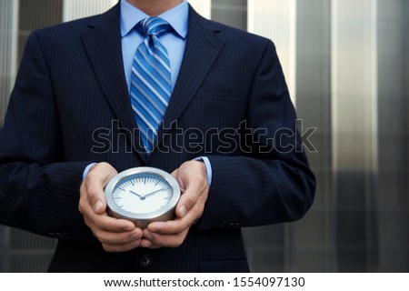 Unrecognizable businessman holding a simple clock standing outdoors in front of modern shiny business background