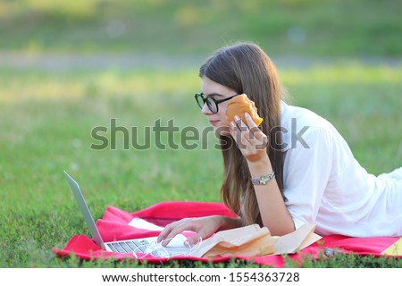 female in the park works behind laptop. beautiful girl with glasses eating fast food in nature and smiling.