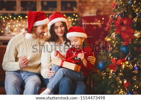 Cute little festive girl openng xmas gifts on Christmas eve with mom and dad, decorated kitchen interior, copy space