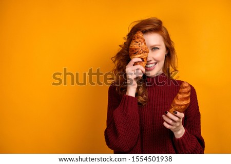 Curly red-haired girl in a burgundy sweater and holds croissants and laughing, covers one half of her face.
