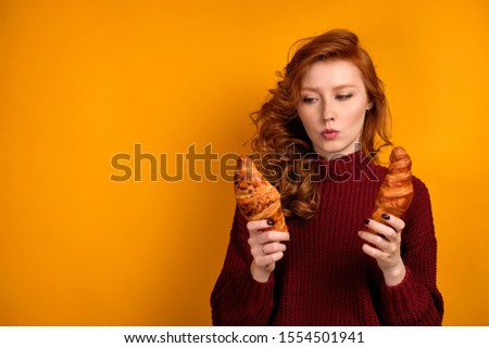 Red-haired curly girl in a burgundy sweater stands on an orange background and chooses between two croissants in her hands.