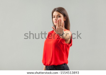 Stop! Portrait of serious beautiful brunette young woman in red shirt standing with stop hand sign gesture and looking at camera. focus on hand. indoor, studio shot, isolated on gray background.