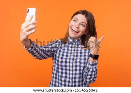 Thumbs up! Portrait of lovely brunette woman with charming smile wearing checkered shirt using cellphone, showing like gesture while making video call. indoor studio shot isolated on orange background