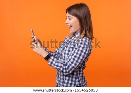 Profile of amazed beautiful brunette woman with charming smile in checkered shirt reading message on cellphone, pleasantly surprised by good news. indoor studio shot isolated on orange background