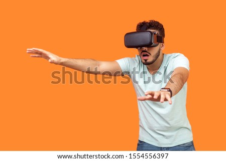 Portrait of scared gamer, brunette man with beard in t-shirt wearing vr glasses, stretching arms forward while playing virtual reality game, frightened face. studio shot isolated on orange background