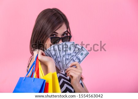 Shopping woman holding shopping bags in one hand and money in other looking at the camera on pink background. Beautiful young shopper smiling happy.