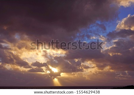 Dark grey clouds with yellow highlights against a blue sky, with one bright yellow sunbeam, and dark ocean, just before the sun sets.

 
