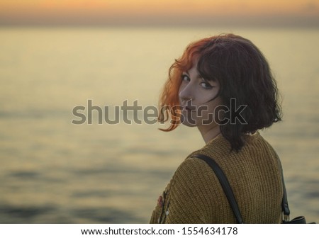 hipster woman with red and black hair looking into the camera with ocean sunset background