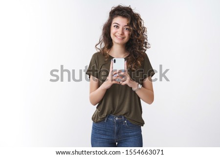 Girl taking your photo. Friendly-looking young cute girlfriend curly-haired holding smartphone capture vertical photograph smiling broadly camera, standing white background shooting friend
