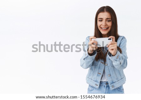 Distressed, intense young attractive woman in denim, holding smartphone horizontally, frowning and grimacing as losing online mobile game battle, playing on telephone, standing white background