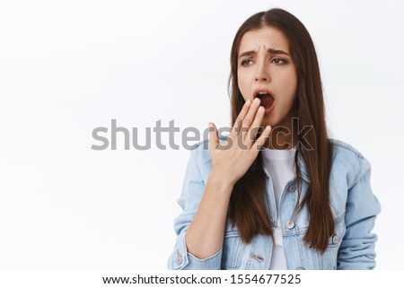 Bored and sleepy young tired brunette girl in denim jacket, frowning look away and yawning, cover opened mouth, need sleep, wake up early morning want cup of coffee, standing white background