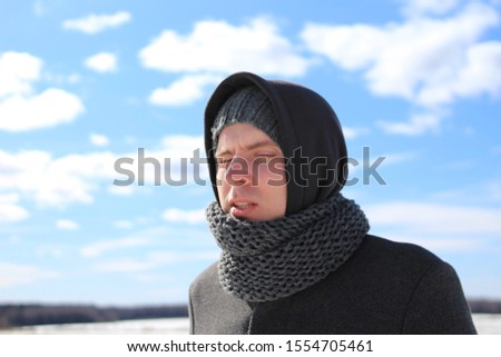 a young man wrapped in a hood, a hat and a scarf, dressed in a coat, is still cold on a clear frosty winter day