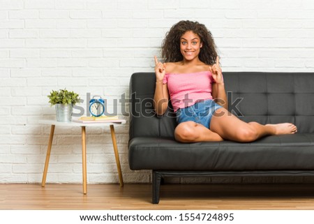 Young african american woman sitting on the sofa indicates with both fore fingers up showing a blank space.