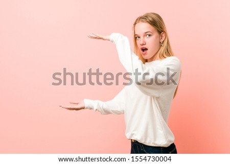 Young blonde teenager woman shocked and amazed holding a copy space between hands.
