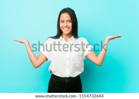 Young hispanic cool woman against a blue wall makes scale with arms, feels happy and confident.