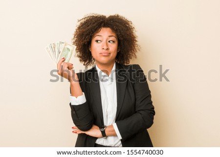 Young african american woman holding dollars looking sideways with doubtful and skeptical expression.