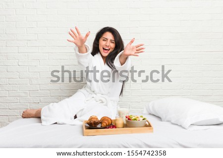 Young curvy woman taking a breakfast on the bed feels confident giving a hug to the camera.