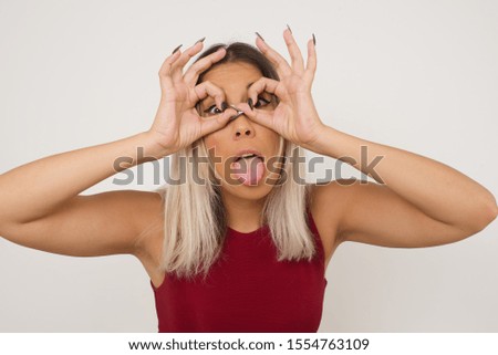 Playful excited teenage female with dark long hair showing Ok gestures with both hands, pretending to wear spectacles, astonished to see something amazing. Emotions, body language and gestures.
