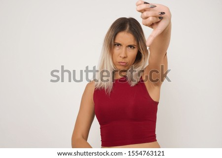 Discontent European woman shows disapproval sign, keeps thumb down, expresses dislike, frowns face in discontent, dressed in white shirt, isolated over gray background. Body language concept.
