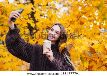 Happy girl in brown sweater is doing selfie using a smartphone and smiling in the autumn park
