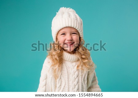 little blonde girl in white winter knitted hat and sweater on blue background isolate, space for text. the child rejoices in winter and snow. winter clothes