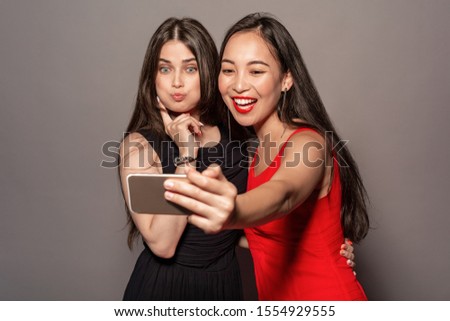 Young women wearing evening dress studio standing hugging isolated on gray background holding smartphone taking selfie picture pouting lips grimacing cute