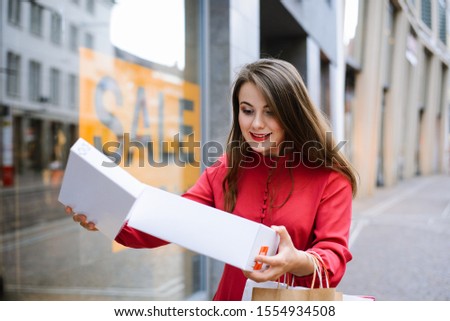 Cheerful caucasian girl in fashionable red dress with new purchase bought while discount period. Sale shopping season, sale label sign sticker in shop window.