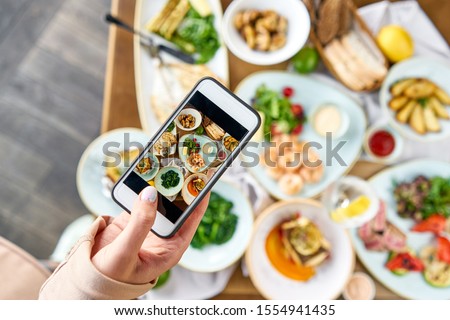 A blogger takes a photo on a smartphone for social networks. Table with Plates seafood BBQ with garnish. Variety of dishes on the table. Restaurant menu.