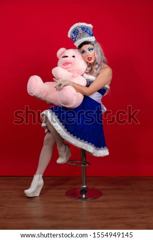 Emotional Snow Maiden in an elegant bright blue suit with a crown posing with a big teddy bear on a red background