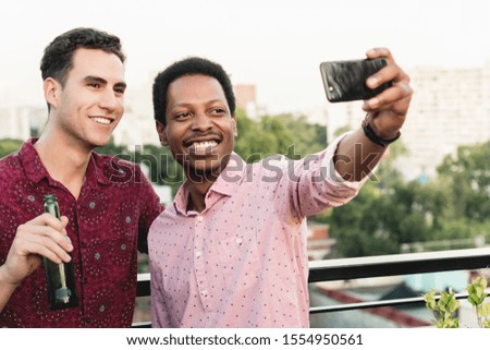 Two male friends taking a selfie while holding beer bottles on the rooftop.