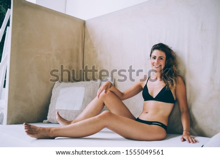 Chilling modern fit female in black bikini sitting on white resort lounge with cushion having vacation and looking at camera