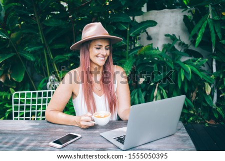 Beautiful female blogger wearing hat and white top at workplace in green garden looking at laptop screen on warm summer day