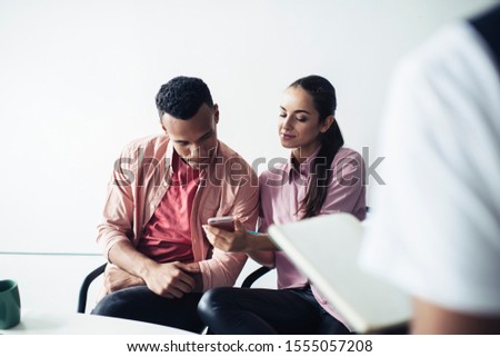 Attractive hipster girl with smartphone gadget in hand reading text in social networks and showing publication to male colleague sitting near, diverse man and woman using smartphone with wireless 4g