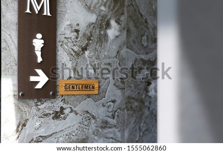 Diagonal view of restroom entrance and focus on white male restroom sign with arrow and gentleman wooden label on gray marble wall with blur foreground