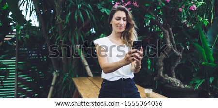 Beautiful woman wearing white shirt and black shorts texting on cellphone while standing at wooden table in cafe decorated with tropical plants and looking at screen