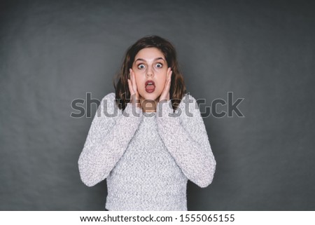 Surprised excited young female wearing sweater looking at camera and touching face with both hands while standing in studio on gray background 