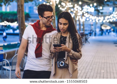 Smiling handsome adult male in casual wear with glasses cuddling beautiful female with smartphone in hands and photo camera on neck while promenading on square