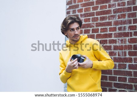 Handsome male photographer wearing sweatshirt standing with camera in hands at coner of brick building and looking for interesting object ready to take picture