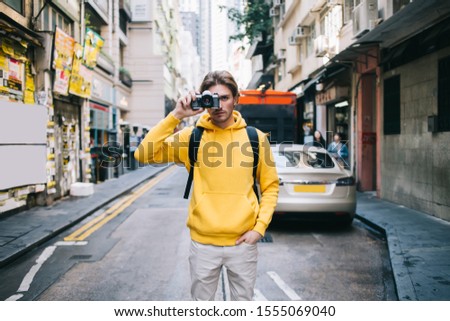 Young male tourist with backpack wearing sweatshirt standing on street side with hand on pocket and taking picture with photo camera 