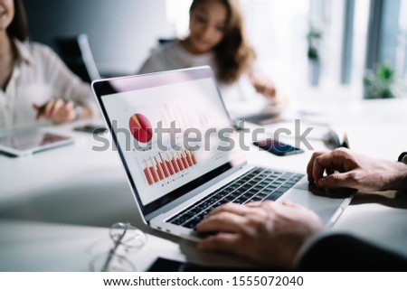 Crop employee using laptop and researching colorful analytical diagrams and statistics while sitting at table with female coworkers on background 