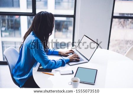High angle side view of ethnic busy woman typing on laptop and using tablet while sitting at table in light office and relishing beverage