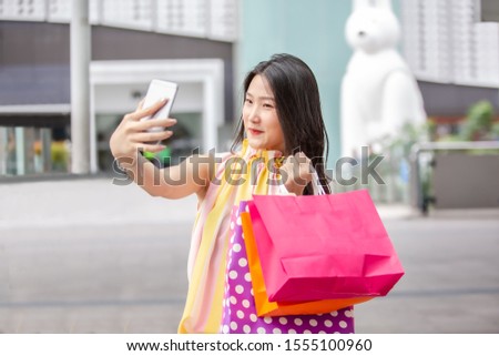 Young beautiful Asian woman holding her smartphone to taking a selfie with her shopping bags in the city street