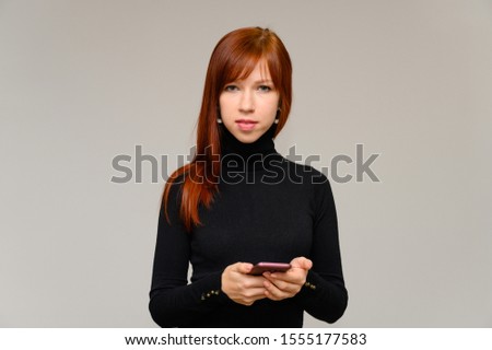 Portrait of a pretty red-haired student girl with long smooth hair on a white background in a black sweater with a phone in her hands. Talking, showing emotions, smiling.