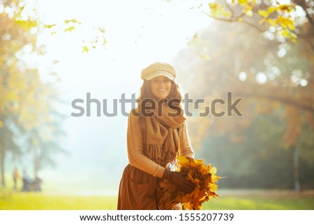 Hello autumn. Portrait of happy young woman in sweater, skirt, hat, gloves and scarf outdoors in the autumn park holding a pile of yellow leaves.