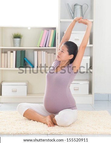 Pregnancy yoga meditation. Full length healthy 8 months pregnant calm Asian woman meditating or doing yoga exercise at home. Relaxation arms stretching pose.