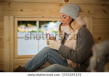 Young woman drinking cup of hot chocolate with marshmallow wearing knitted warm hat and scarf. Girl sitting next to window in traditional Finnish wooden hut on Christmas market in Helsinki, Finland