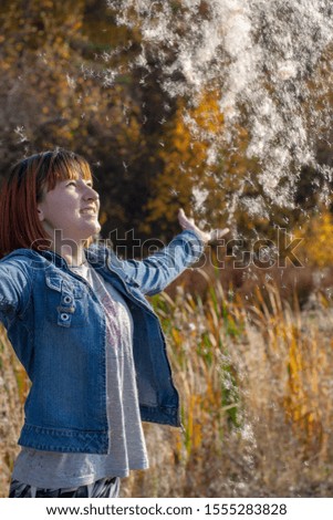 A teenage girl with red hair throws up fluff from a cattail and he falls down. Autumn forest and grass on a blurred background.