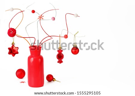 Funny Christmas still life with red vase, colorful decorative twigs and red Christmas decorations on a white background. Close up. Monochrome. Free space on the right. Humorous picture.

