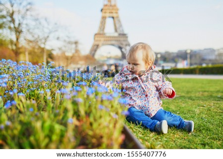 One year old girl sitting on the grass with blue flowers and Eiffel tower in the background. Toddler looking at flowers on a spring day in Paris, France. Adorable little kid exploring nature